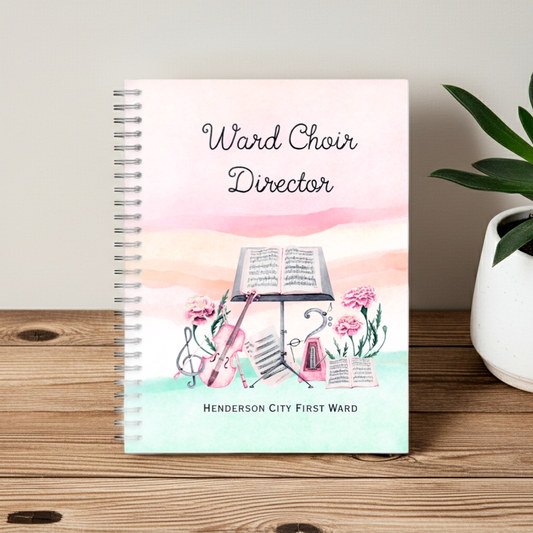 A spiral-bound notebook standing upright on a wooden surface, featuring a pastel cover design with the text 'Ward Choir Director' and 'HENDERSON CITY FIRST WARD,' plus an illustration of sheet music, flowers, and musical instruments. A potted plant is visible at the right edge.